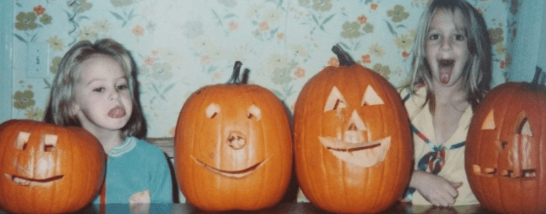 Two young girls sticking their tongues out with four jack-o-lanterns on the table.