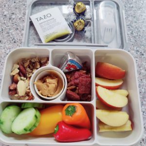 Bento box with cucumber, peppers, apples, peanut butter, cheese, beef jerky, trail mix and chocolate.