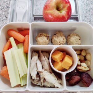 Bento box with tahini medjool date cookies, turkey, cheese, macadamia nuts, carrots, celery, dried apricots and an apple.