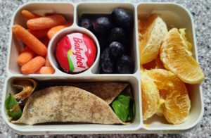 Bento box with peanut butter and jelly wrap, cheese, carrot, grapes and orange slices.